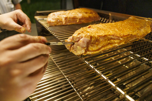 Ein Mann sticht ein Thermometer in ein mariniertes Schweinefilet, das auf einem Grillrost liegt. Ein weiteres Filet liegt im Hintergrund auf einem zweiten Rost. Die Hände des Mannes sind mit Marinade bedeckt.
